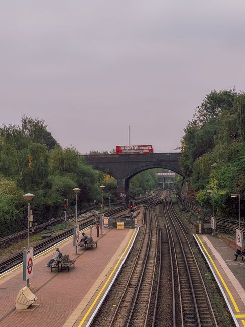 View of Charlton Station platform with three wooden benches, two of which are occupied by individuals sitting and waiting, alongside a stationary bicycle rack. The platform is paved with red-brown bri